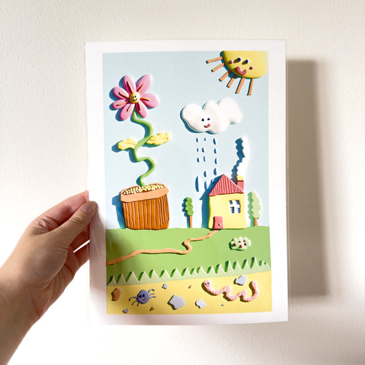 Hand holding a colorful 3D giclée art print with a house, flower, and rain cloud on a white background