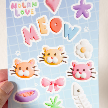 A close up of a hand holding a sheet of colorful stickers with cat-themed designs held by a hand against a light background.