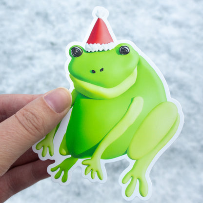 Green frog sticker with a Santa hat held by a hand against a light background