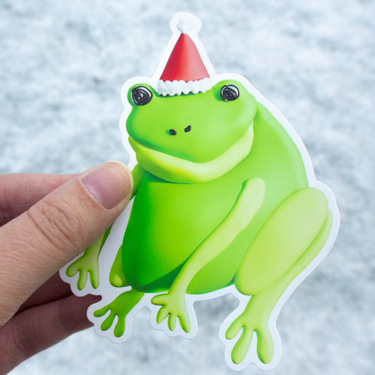 Green frog sticker with a Santa hat held by a hand against a light background