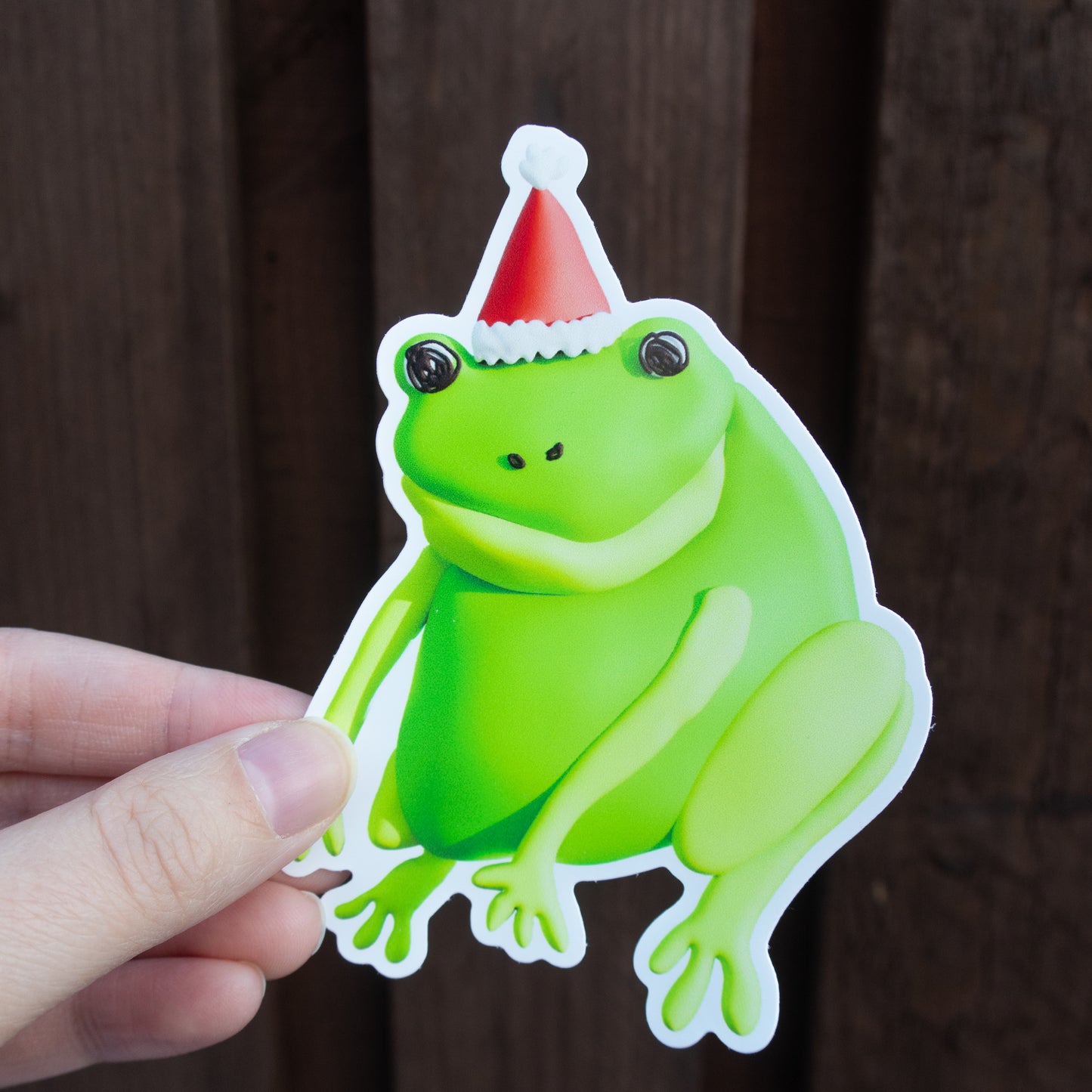 Green frog sticker with a red and white hat held by a hand against a wooden background