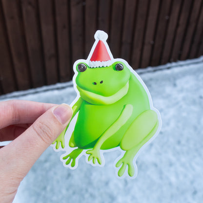 Green frog sticker with a red and white hat held by a hand against a wooden fence background.