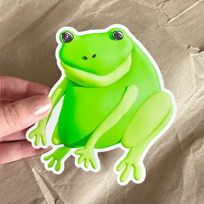Hand holding a large green frog sticker against a brown paper background.
