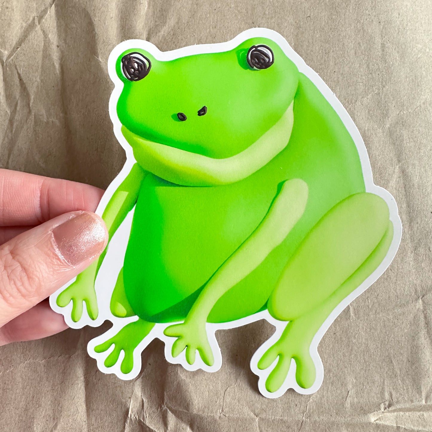 Hand holding a large green frog sticker against a brown paper background.