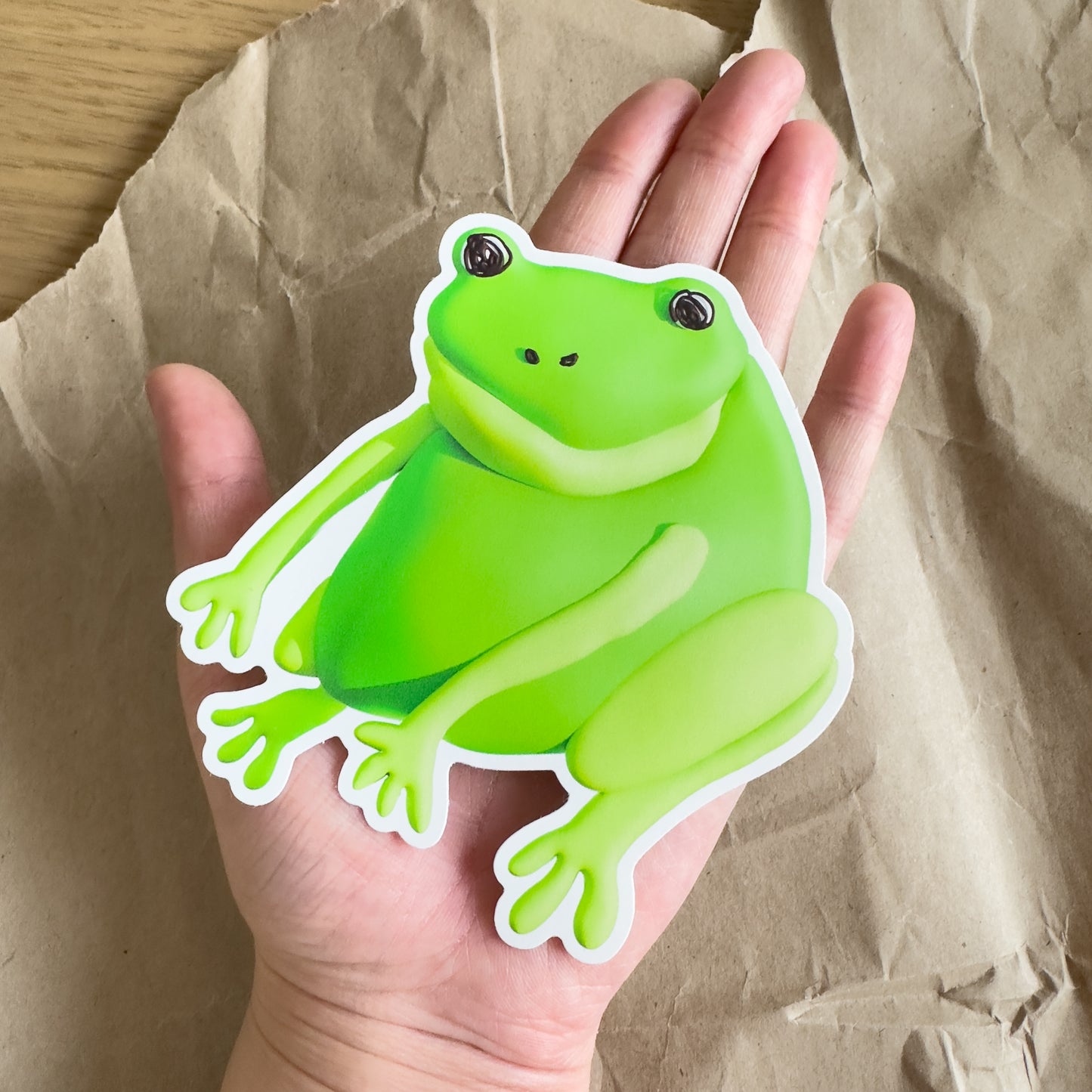 An outstretched hand holding a large green frog sticker against a brown paper background.