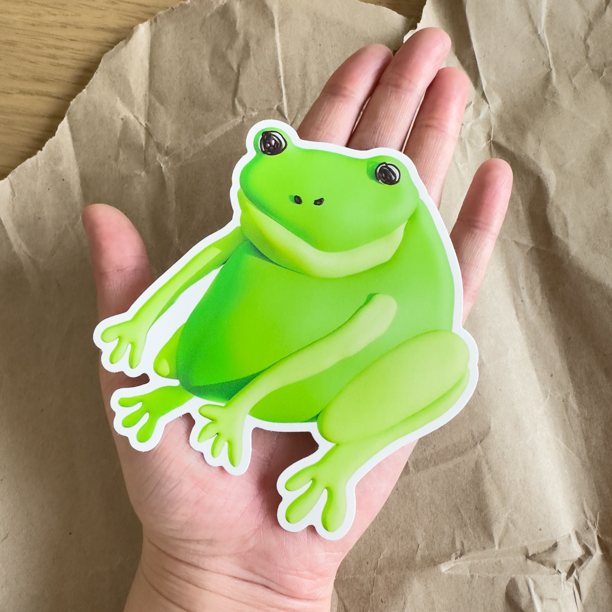 An outstretched hand holding a large green frog sticker against a brown paper background.