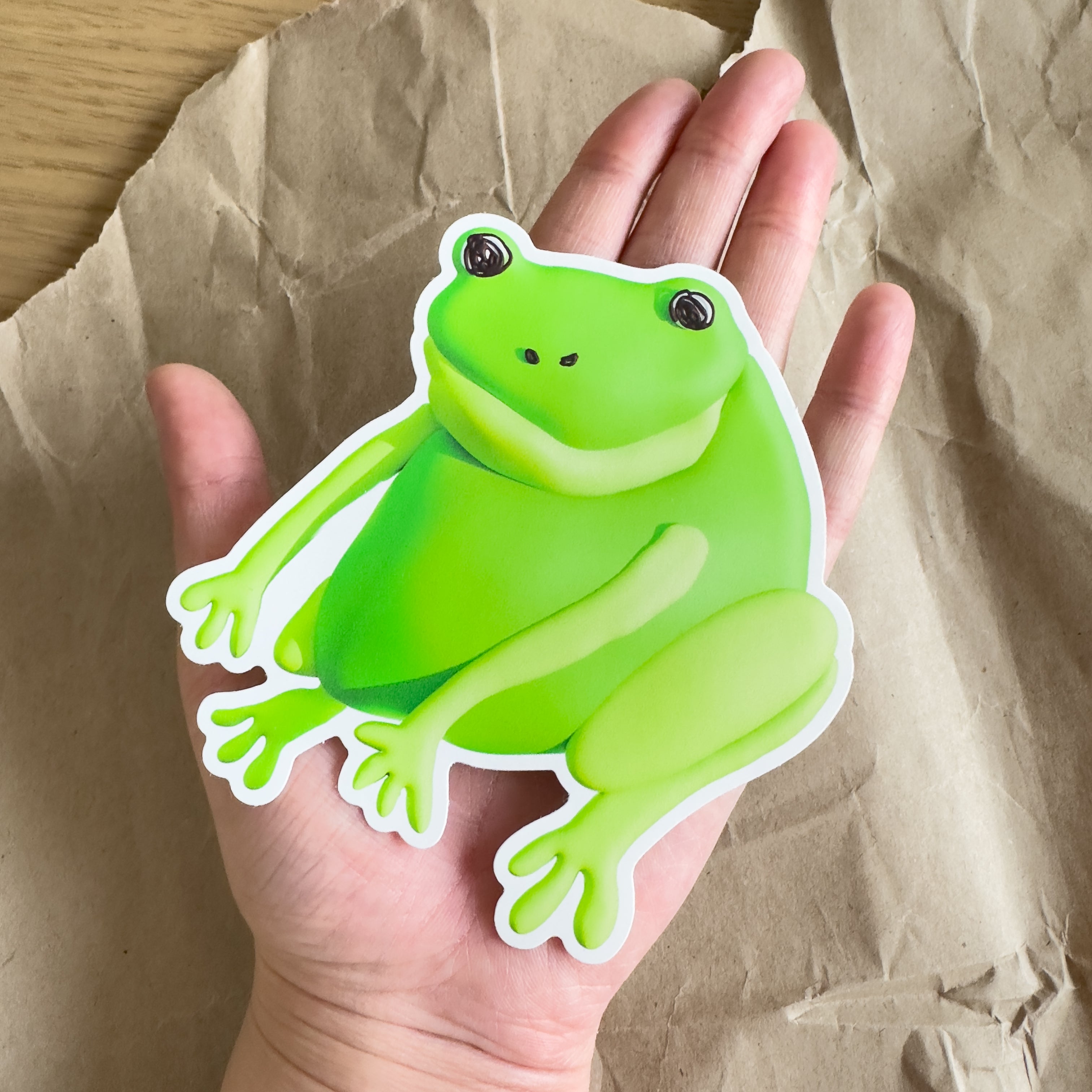 An outstretched hand holding a large green frog sticker against a brown paper background.