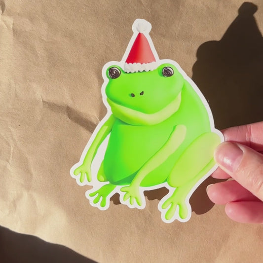 Video of a green frog sticker with a Santa hat held by a hand against a light brown paper background