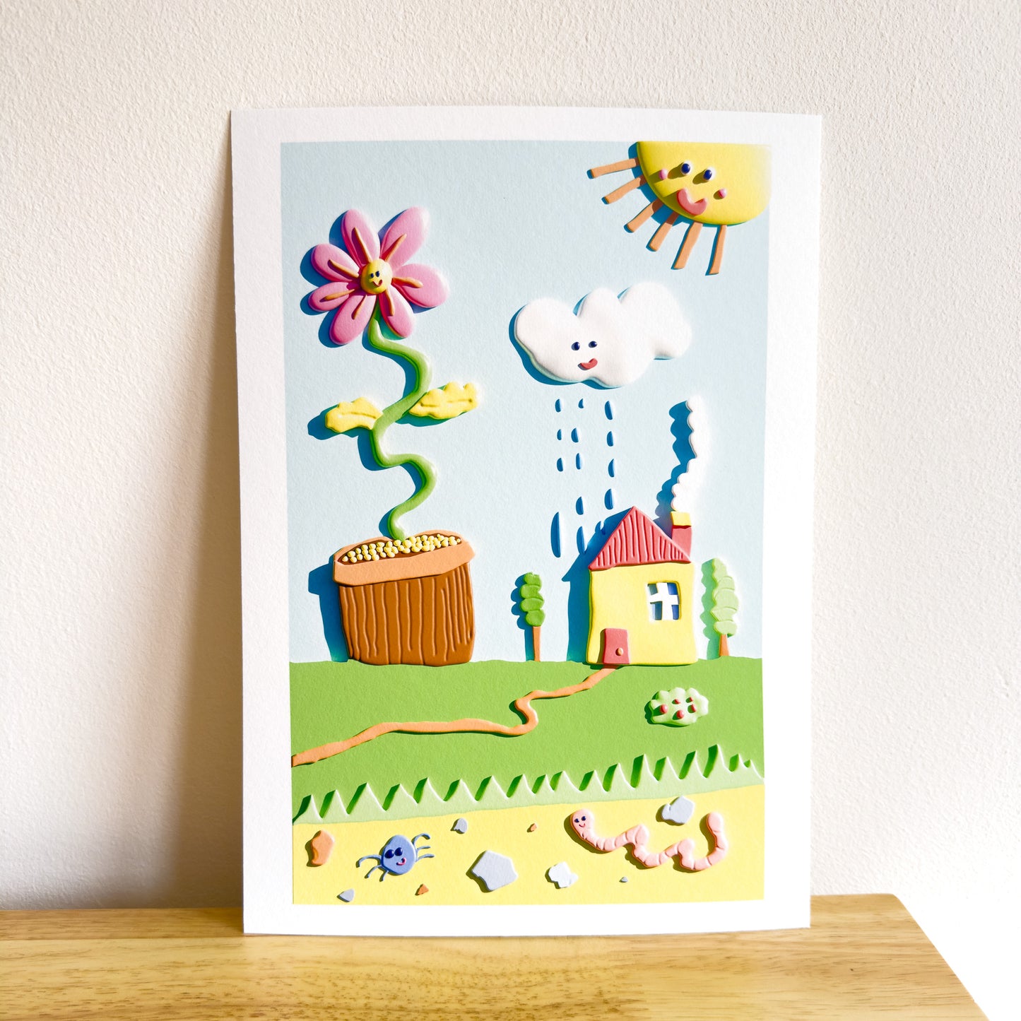Hand holding a colorful 3D giclée art print with a house, flower, and rain cloud on a white background